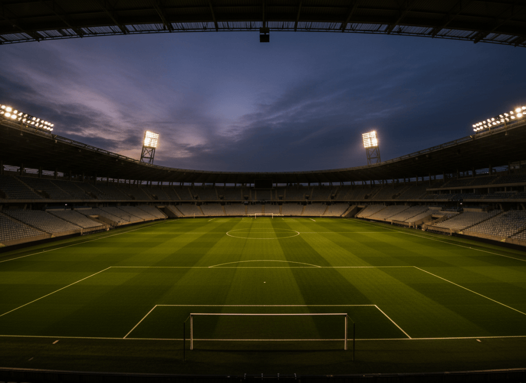 A dramatic, empty football stadium at dusk or night. No people, no players. Grand curved stands, floodlights casting a warm glow over a lush green pitch. Cinematic wide-angle perspective from pitch level. Dark, moody atmosphere with deep shadows and vivid green turf. Professional sports photography style. High contrast, editorial feel.